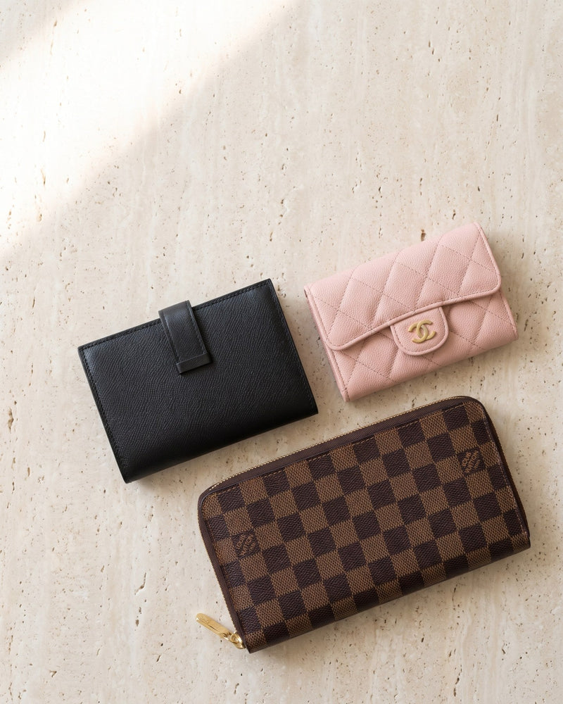 A black Hermès Béarn wallet, a pink Chanel card holder and a Louis Vuitton Damier zip wallet on travertine