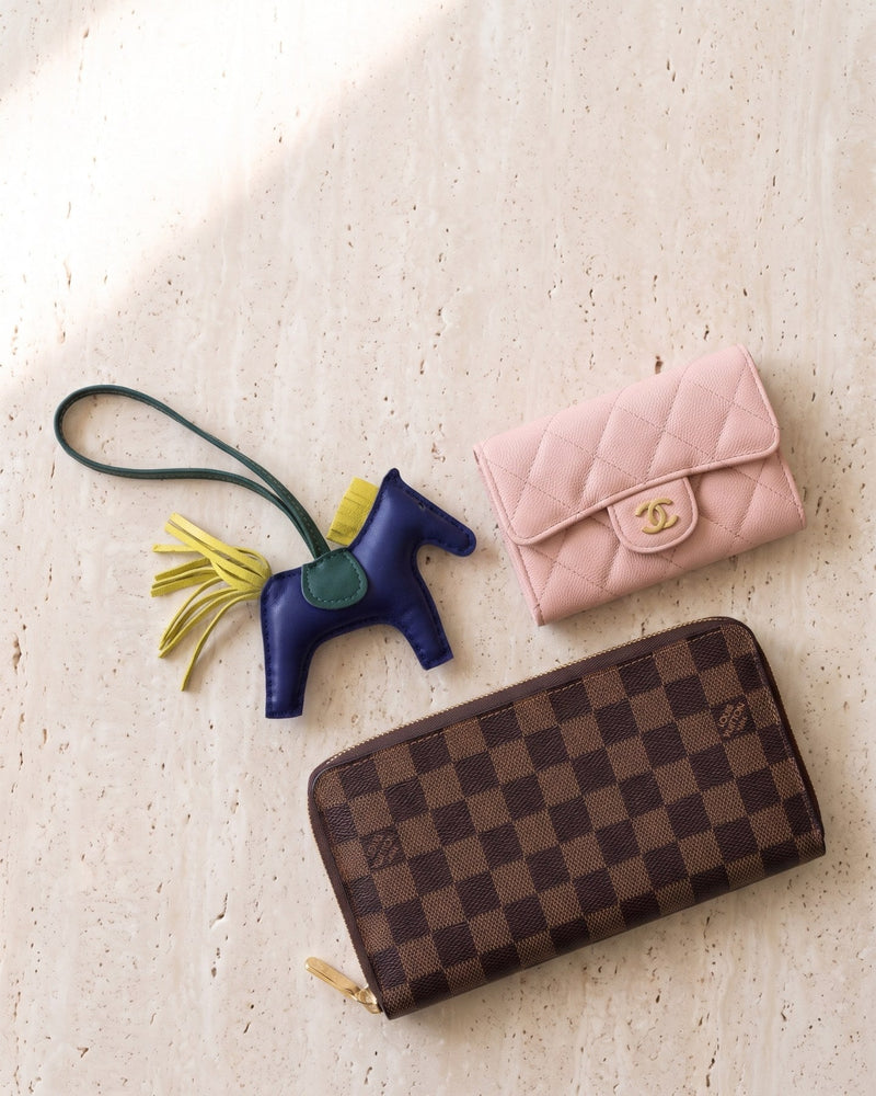 A pink Chanel card holder, a Hermès Rodeo bag charm and a Louis Vuitton Damier zip wallet on travertine